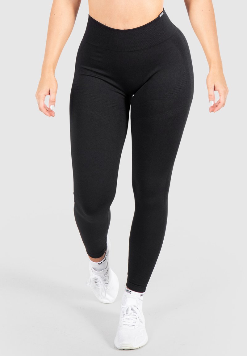 Leggings neri a vita alta realizzati in tessuto elasticizzato, caratterizzati da una texture liscia e da un design aderente, abbinati a scarpe da ginnastica bianche.