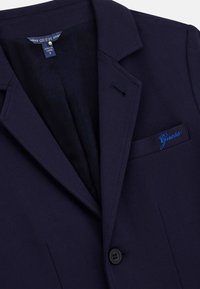 Marinblå blazer med slagkrage, enknappsstängning och en vänster bröstficka med en blå broderad "Guess"-logotyp.