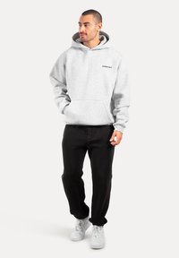 Felpa grigia con cappuccio e tasca a marsupio sul fronte, vestibilità comoda e dettagli in logo nero. Abbinata a pantaloni neri e sneakers bianche.