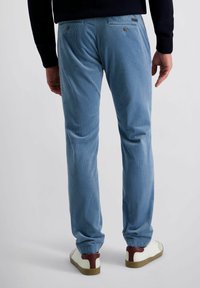 Man in lichtblauwe corduroy broek en witte sneakers met bruine hielaccenten, staand tegen een effen lichte achtergrond.