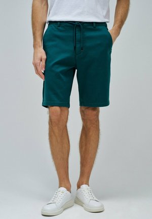 Pantalones cortos de algodón en color azul verdoso con cintura elástica y cordón ajustable; cuentan con bolsillos laterales y dobladillo recto. Combinados con zapatillas blancas.