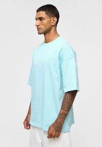 T-shirt bleu clair à manches courtes en coton, avec une coupe décontractée et un col rond. Détail remarquable : motif de tatouage imprimé sur le bras.