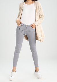 Licht beige gebreide cardigan over een witte top, gecombineerd met grijze skinny jeans en witte sneakers. De outfit heeft een ontspannen maar toch aansluitende silhoutte.
