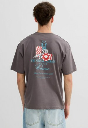 Hombre con camiseta gris con un gráfico colorido de ficha de casino y texto "The Grand Mirage Hotel & Casino" en la espalda, combinado con jeans azul claro.