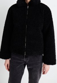 Person wearing a black textured zip-up jacket with long sleeves and dark gray jeans, standing against a plain background.