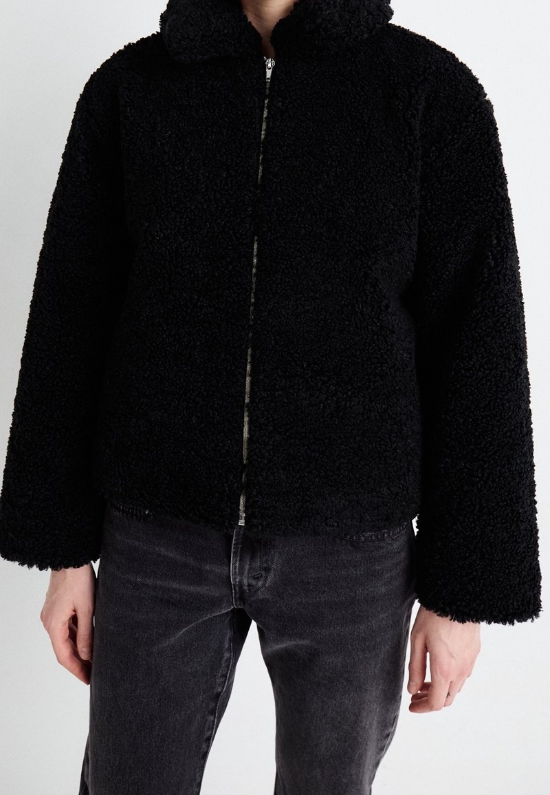 Person wearing a black textured zip-up jacket with long sleeves and dark gray jeans, standing against a plain background.