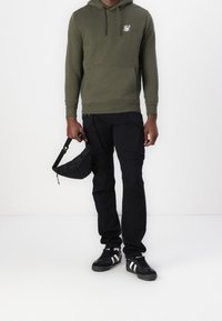 Sudadera verde oliva con bolsillo frontal y capucha ajustable. Pantalones cargo negros con múltiples bolsillos. Zapatillas negras con rayas blancas.