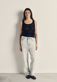Young woman standing against white wall wearing black sleeveless top, light blue jeans, and black woven sandals.