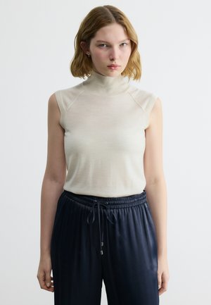 Ženska s kratkimi blond lasmi, oblečena v brezrokavnik-bež pulover z visokim ovratnikom in temno modre hlače z vrvico, stoji pred enobarvnim belim ozadjem.