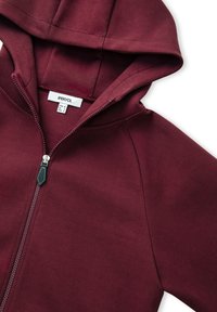Sudadera con cremallera en color burdeos, con capucha, textura suave y etiqueta interior con instrucciones de cuidado. La cremallera tiene una lengüeta en contraste.