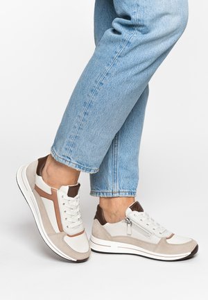 Person wearing blue jeans and beige and brown sneakers with white soles, standing with one foot slightly raised.