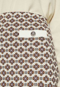 Gros plan d’un tissu à motif avec des formes florales géométriques répétées en rose, orange et noir sur fond blanc, avec une poche côtelée blanche et un bouton argenté.