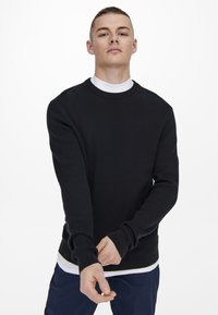 Pull noir à côtes avec des manches longues, col rond et poignets ajustés ; superposé à une chemise à col roulé blanche, en tissu texturé.