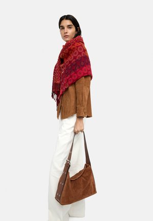 Kobieta w brązowej kurtce i czerwonym szaliku w wzory, trzymająca brązową zamszową torebkę, stojąca na jednolitym tle.
