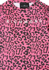 Różowa koszulka krótkim rękawem z czarnym nadrukiem w panterkę. Logo "RICHMOND" w białym kolorze na przodzie. Dopasowany krój z falbaniastymi akcentami na ramionach.