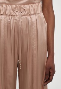 Pantalons larges en satin rose clair avec taille élastique, comportant des plis et des accents en métal sur les extrémités des cordons de serrage.