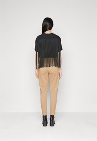Černé tričko s třásněmi na zádech, v kombinaci s béžovými kalhotami a černými botami na vysokém podpatku. Minimalistický design na světlém pozadí.