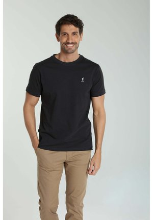 Homme souriant portant un t-shirt noir à manches courtes avec un petit logo et un pantalon beige, debout avec une main dans la poche, sur un fond uni.