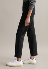 Schwarze, maßgeschneiderte Hosen mit glatter Textur und geradem Schnitt, kombiniert mit weißen Sneakern, die dezente Markenakzente und eine kontrastierende Sohle aufweisen.