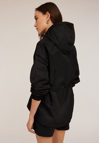 Svart huva jacka med elastiska ärmar, lättviktsmaterial och en lös passform. Har en dra-in midja och en slät, matt struktur.