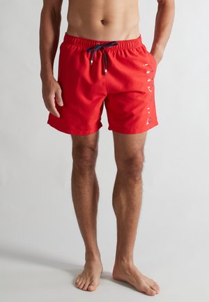 Man wearing bright red swim shorts with black drawstring and white text logo on right thigh, standing barefoot against plain background.