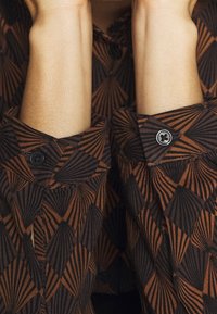 Brown and black patterned shirt with geometric designs, featuring buttons and rolled-up cuffs. Material texture appears smooth and lightweight.