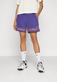 Lila sportnadrág narancssárga csíkokkal, amelyen Nike logo található. Fehér zoknival és fekete-fehér tornacipővel párosítva, sportos megjelenésért.