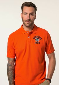 Světle oranžová polo košile vyrobená z texturované tkaniny. Obsahuje dva knoflíky a černě vyšitý logo "Vancouver 1880" na hrudi.