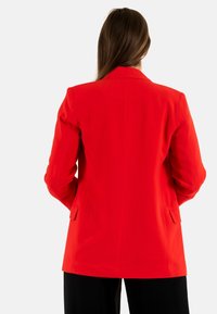 Blazer rojo con solapa de muesca, corte ajustado y bolsillos laterales. Tejido suave con un diseño estructurado; vista desde la parte trasera. Pantalones negros visibles.