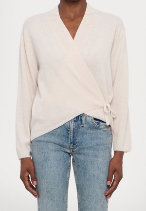 Personne portant un pull cache-cœur beige clair noué sur le côté et un jean bleu taille haute, coupé au milieu du torse avec les bras détendus.