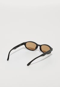 Black sunglasses with an oval frame, brown tinted lenses, and smooth, glossy plastic arms. Notable details include subtle branding on the side.
