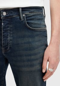 Jeans de mezclilla azul oscuro con un corte ajustado, que presentan un diseño clásico de cinco bolsillos, un ligero desvanecimiento y un cierre de botón plateado.