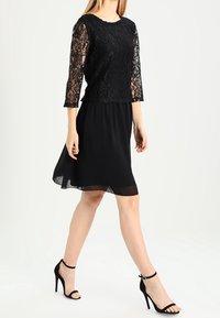 Kvinna som går i en svart knälång klänning med spetsärmar och svarta högklackade sandaler mot en enfärgad vit bakgrund.