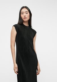 Fekete bordás ruha magas nyakú és kis ujjas megoldással. Az anyag texturált megjelenésű, sima és elegáns sziluettet kínál.