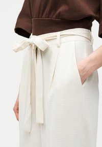 Κοντινή λήψη κρεμ παντελονιού ψηλής μέσης με δεμένη υφασμάτινη ζώνη και πλαϊνή τσέπη, συνδυασμένο με καφέ τοπ. Το χέρι ακουμπά στην τσέπη.