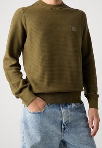 Sweatshirt en tricot vert olive avec un col rond, surface texturée et poignets et ourlet côtelés. Présente un petit patch de logo de la marque.