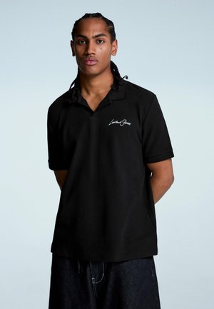 Young man with braided hair wears black polo shirt with white logo, standing against plain light background with hands behind back.