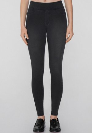 Jeggings noirs avec une taille haute, présentant une texture lisse, de fausses poches et une silhouette ajustée. Associés à des mocassins noirs.