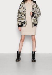 Giubbotto bomber camouflage con dettagli neri, abito in maglia beige testurizzata, e stivali neri con lacci. Abbigliamento aderente e stratificato.