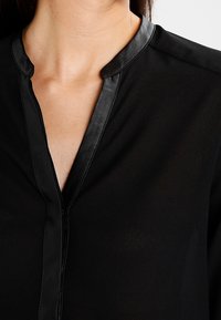 Femme portant un blouse noire avec un décolleté en V et une bordure en simili cuir brillant le long du col et de la patte de boutonnage.