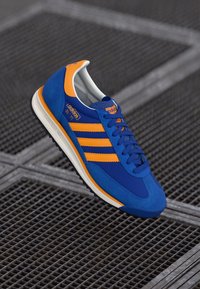 Blue suede sneaker with orange accents, featuring three stripes, round toe, and a textured sole. Model name "SL 72" is visible on the tongue.
