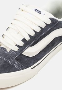 Zapatillas de ante gris con cordones blancos, que cuentan con una franja lateral blanca en contraste, suela de goma y un detallado de costuras.
