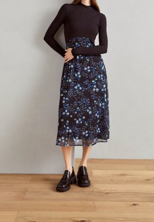 Kobieta w czarnej bluzce z długim rękawem, z wysokim stanem, czarnej kwiecistej midi spódnicy i czarnych mokasynach na platformie stoi na drewnianej podłodze.