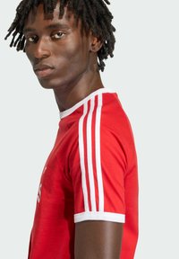 Červené športové tričko s bielymi akcentmi, ktoré má na každom pleci tri biele pruhy a okrúhly výstrih. Mäkká, mierne priliehajúca látka.