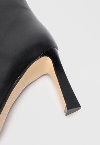 Chaussure noire en cuir à talon haut avec un bout pointu et un talon carré. L'intérieur est beige, mettant en valeur des textures lisses et un design épuré.