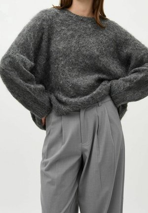 Femme portant un pull ample et duveteux gris foncé à manches longues et un pantalon taille haute plissé gris clair, sur un fond uni.