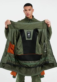 Grüne wasserdichte Jacke mit Netzfutter, Innentaschen und orangefarbenen Akzenten. Verfügt über ein auffälliges Markenlabel und verstellbare Riemen.