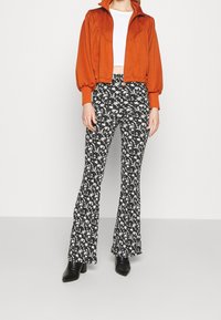 Chaqueta corta naranja con mangas abullonadas, combinada con pantalones acampanados negros que presentan un patrón floral blanco. La modelo lleva botas de tacón negras.