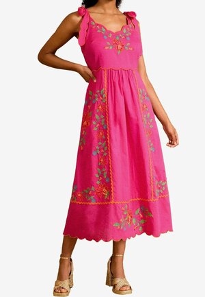 Femme portant une robe midi rose vif avec broderie florale et bords festonnés, associée à des sandales à talons hauts dorées.