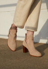 Bottines en daim beige avec un talon épais et des détails découpés sur les côtés. Elles présentent un talon texturé imitant le bois et des accents de brides simples.